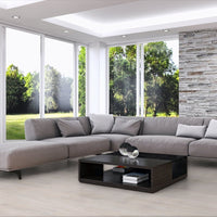 A modern L-shaped sectional sofa in light gray fabric, styled in a contemporary living room with a black coffee table, a side table, a stone accent wall, and large windows overlooking a green lawn.