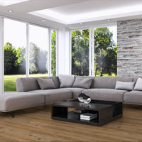 A modern light gray sectional sofa with matching throw pillows arranged in an open U-shape in a bright living room. The room features a light wood floor, a black coffee table, a textured stone accent wall, and large windows looking out onto a green lawn.