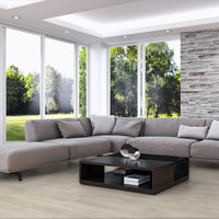 A modern L-shaped sectional sofa in light gray fabric, styled in a contemporary living room with a black coffee table, a side table, a stone accent wall, and large windows overlooking a green lawn.
