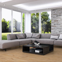 A modern gray sectional sofa with matching throw pillows arranged in an open U-shape in a bright living room. The room features a light wood floor, a black coffee table, a textured stone accent wall, and large windows looking out onto a green lawn.